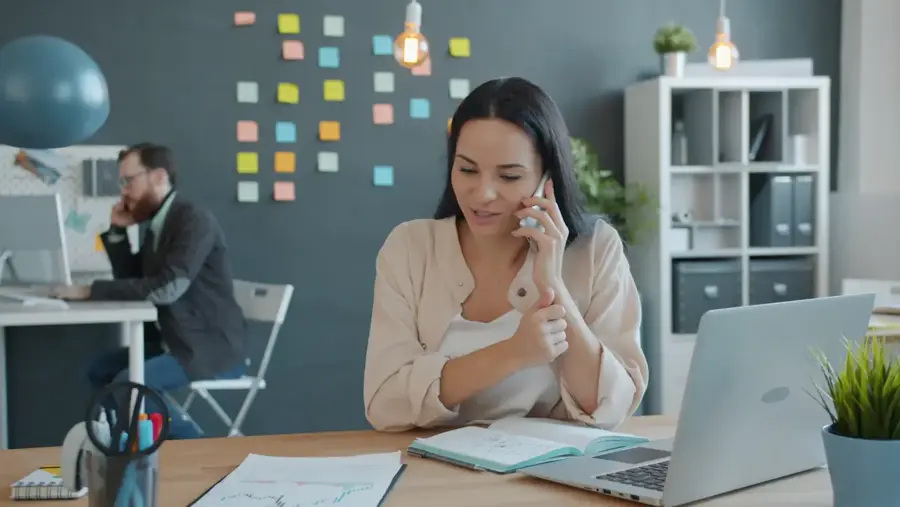 A woman at a desk talking on the phone, notebook open beside her laptop